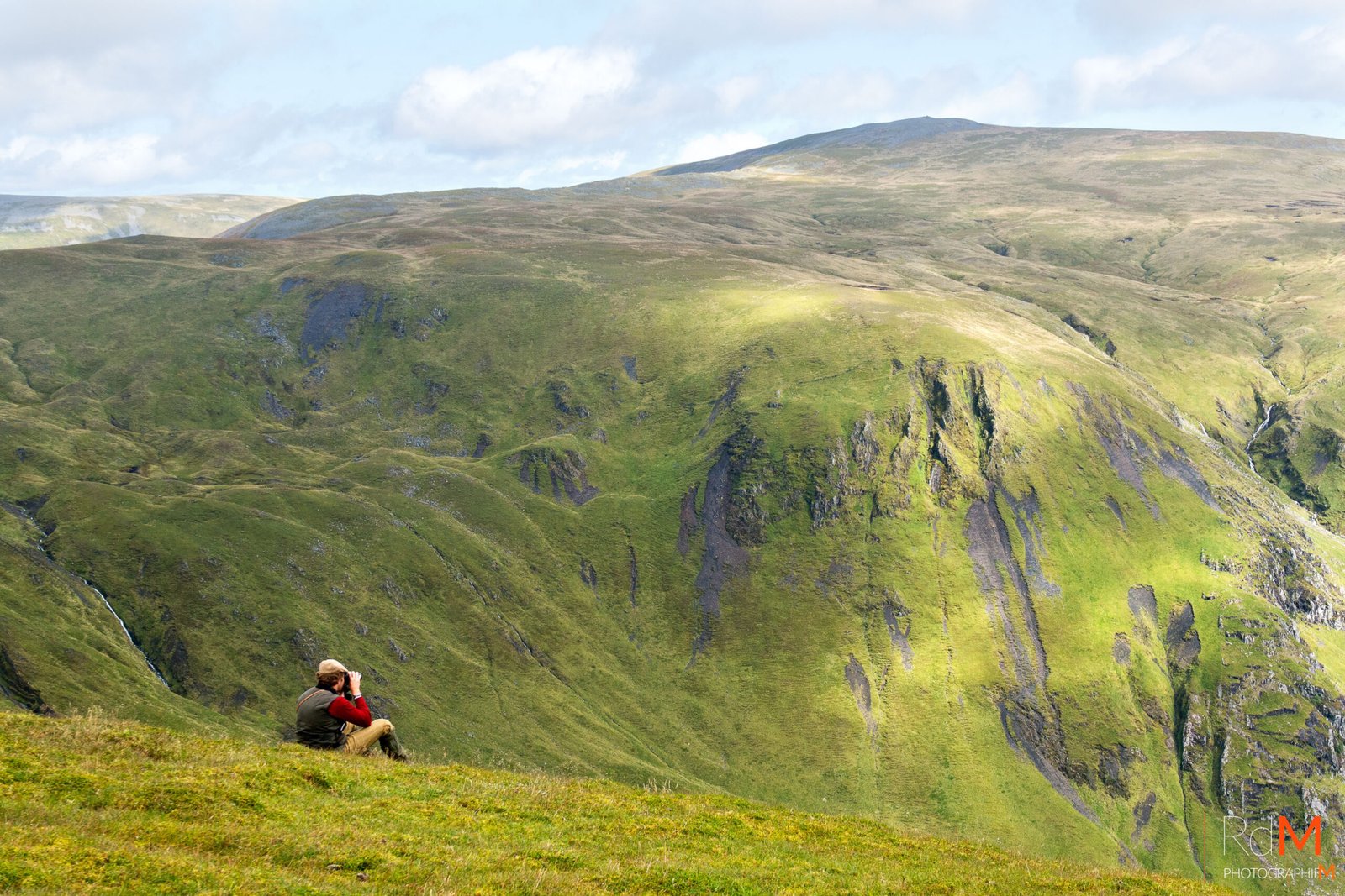 Personne assise sur une colline verdoyante, observant des montagnes et vallées au loin.